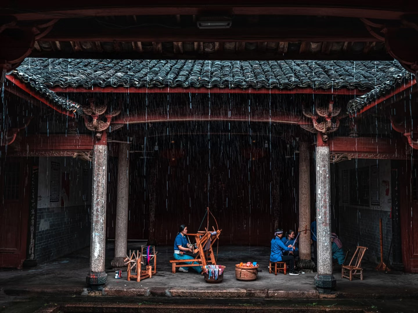 Traditional scene with people using a loom under a tiled roof during rain.